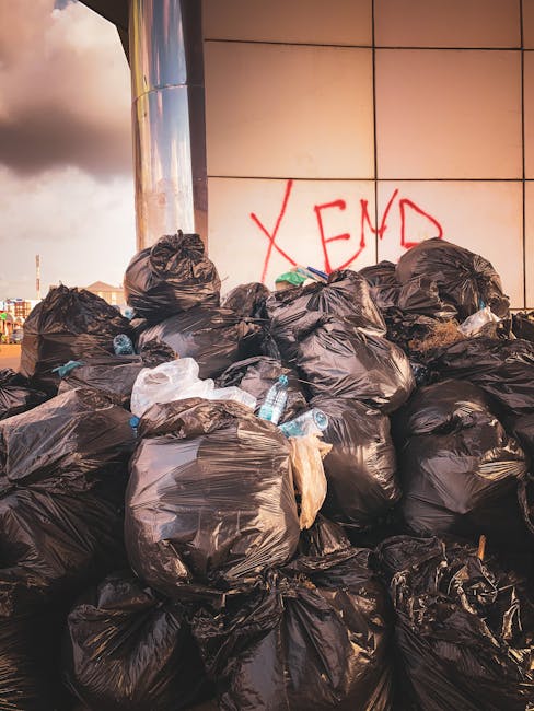 A large accumulation of black plastic rubbish bags stacked against a modern building with metallic and tiled exterior panels. The bags are tightly packed and vary in size, with some appearing to be slightly torn or bulging. Several transparent plastic water bottles and other small waste items are visible protruding from or resting on top of the bags. Behind the rubbish, the building's wall features a bold red spray-painted graffiti with the word 'XEND' partially visible. The scene is outdoors, with a cloudy sky and distant urban structures faintly visible in the background. The image highlights a scenario of improper waste disposal in an urban environment, which could relate to any form of private rubbish collection or non-authority waste handling, aligning with themes of independent rubbish removal services in Fulham.