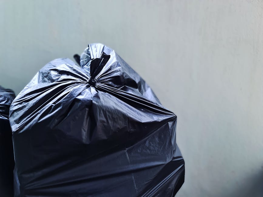 Close-up view of multiple large black plastic rubbish bags tightly sealed with twist ties, resting on a surface against a plain light-grey wall. The bags appear to be made of shiny, slightly crinkled plastic material, reflecting some ambient light. The foreground bag is prominent, occupying most of the image, with others stacked slightly behind and to the left, indicating they are part of the same waste collection or disposal activity. The setting suggests an indoor or outdoor area dedicated to rubbish accumulation, possibly in preparation for private collection or removal service, as commonly used by companies like Rubbish Removal Fulham. The background is unobtrusive, emphasizing the presence of the waste bags, which are associated with activities of rubbish clearance, recycling, or alternative waste handling solutions outside of standard municipal collection. The overall scene emphasizes the practical aspect of waste management in a tidy and organized manner.