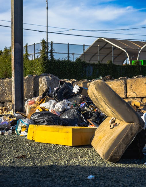 A large accumulation of black plastic rubbish bags stacked against a modern building with metallic and tiled exterior panels. The bags are tightly packed and vary in size, with some appearing to be slightly torn or bulging. Several transparent plastic water bottles and other small waste items are visible protruding from or resting on top of the bags. Behind the rubbish, the building's wall features a bold red spray-painted graffiti with the word 'XEND' partially visible. The scene is outdoors, with a cloudy sky and distant urban structures faintly visible in the background. The image highlights a scenario of improper waste disposal in an urban environment, which could relate to any form of private rubbish collection or non-authority waste handling, aligning with themes of independent rubbish removal services in Fulham.