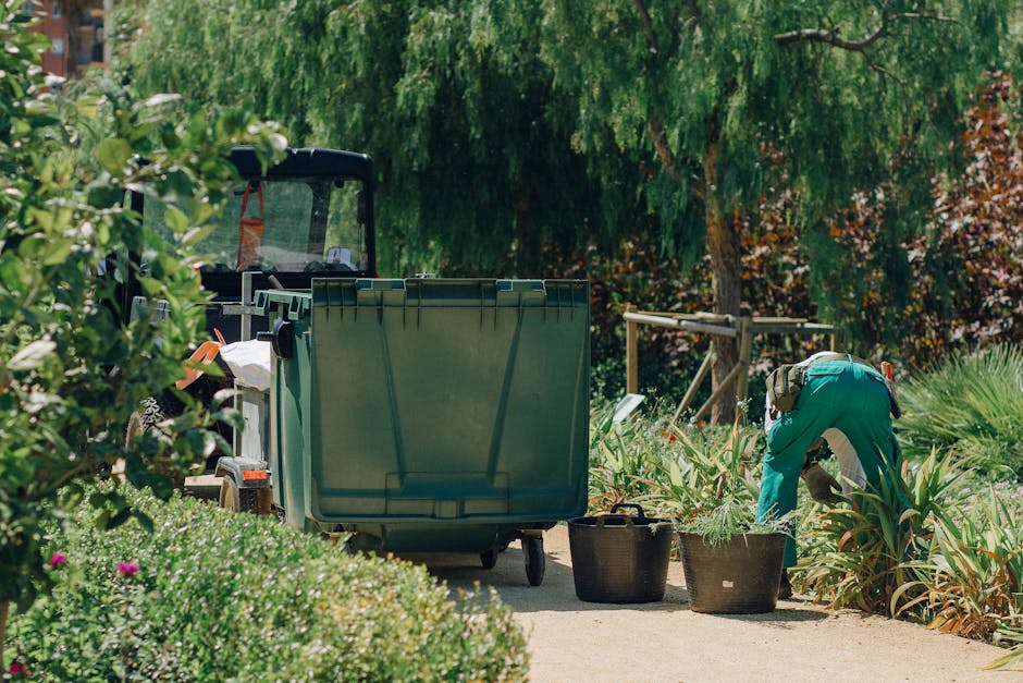 A worker dressed in a teal uniform and protective gloves is bent over, handling plants or garden debris in a lush outdoor garden setting. The scene features a variety of greenery, including tall bushes and flowering plants with pink blossoms, alongside a pathway made of concrete or stone. In the background, a large green company wheelie bin is positioned on the pathway, partially obscuring a small utility vehicle or trailer with a black protective cover. The background includes several mature trees with dense foliage, casting shade over the area. Nearby, a black basket or container is placed on the ground, filled with garden waste such as leaves or branches. The environment suggests an organized garden clearance or gardening activity, with professional waste removal services provided by Rubbish Removal Fulham, specializing in garden and general rubbish clearance, utilizing on-site waste handling options and private disposal methods.