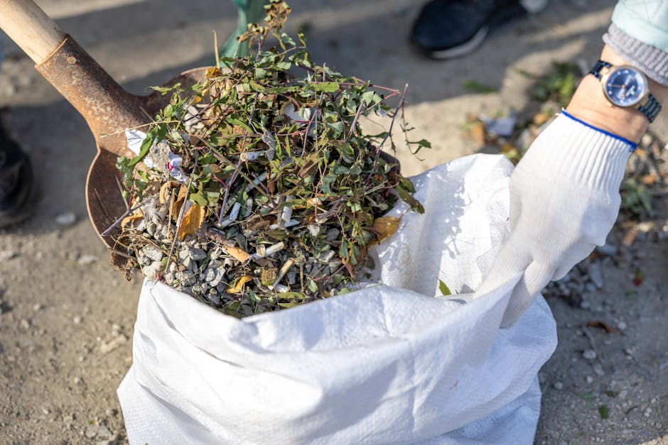 A person wearing a white glove is holding a large white sack filled with garden debris, including small branches, leaves, and twigs with brown and green hues. A metal-handled shovel is positioned above the sack, with its blade partially embedded in the contents, suggesting active collection or loading. The scene is outdoors on a paved or compacted surface, with natural lighting illuminating the debris and surroundings. The background shows a blurred view of the ground and a hint of a person's shoe, indicating an environment where garden clearance or waste removal is taking place. This image reflects the process of garden rubbish collection, which is often handled through private waste removal services, such as those offered by Rubbish Removal Fulham, specializing in efficient clearance of garden waste and debris.