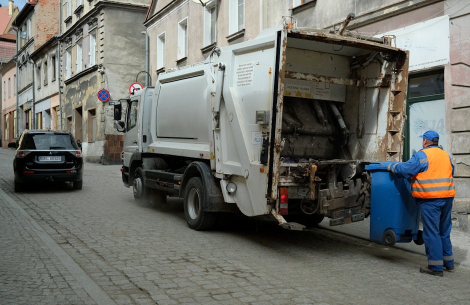 A sanitation worker dressed in a high-visibility orange vest and blue uniform is standing on a cobblestone street, actively placing a blue waste bin into a large, white rubbish collection truck parked adjacent to a row of older, multi-story buildings with weathered facades. The truck, with its rear open to reveal a rusty, metallic interior designed for compacting and collecting waste, dominates the foreground. The truck's exterior shows signs of use, with rust and grime on the inner panels, and the surrounding environment includes a parked black car further down the narrow street, along with street signs indicating parking restrictions. The scene is set during daylight hours with natural lighting highlighting the textures of the cobblestones, the truck's surfaces, and the worn walls of nearby buildings, exemplifying an on-site waste removal process consistent with private rubbish collection services, such as those offered by Rubbish Removal Fulham, in the context of alternative disposal methods for residential waste.