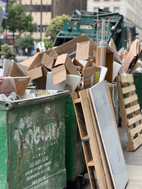 A large open green waste bin filled with a variety of discarded cardboard boxes, some flattened and some still in their original shapes, alongside a wooden pallet leaning against the side. The cardboard boxes are made of corrugated material with visible fluted textures and in different shades of brown and white. In the background, there is a busy urban street scene with a green waste collection truck partially visible and other piles of mixed waste materials. The scene appears to be an area designated for rubbish collection or clearance, possibly managed by a private waste removal service such as Rubbish Removal Fulham, indicated by the context of alternative waste handling methods. Overhead, there are trees with green foliage and surrounding multi-storey buildings, suggesting an outdoor setting in a city environment, with natural daylight illuminating the scene. The arrangement of waste materials and presence of collection equipment reflect a typical site for on-site rubbish clearance or independent disposal activities.