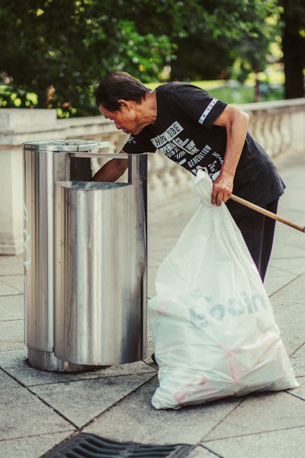 A middle-aged man with short dark hair, dressed in a black T-shirt with white text and dark trousers, is actively disposing of a large white waste bag into a public metal rubbish bin situated on a paved sidewalk. The bin has a cylindrical shape with a reflective, stainless steel finish and an open-top design. The man is holding a wooden stick or litter picker in his right hand, while his left hand guides the waste bag into the bin. The surrounding environment features a stone balustrade and lush green trees, indicating a park or urban outdoor setting. The scene is illuminated by natural daylight, highlighting the textures of the pavement, the polished surface of the bin, and the features of the man’s clothing. This image exemplifies the process of private or independent waste disposal in a public space, aligning with services offered by rubbish removal companies like Rubbish Removal Fulham for efficient on-site clearance and rubbish collection.