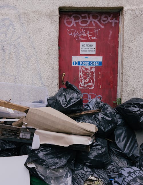 A collection of black garbage bags and loose cardboard boxes piled against a rough, beige exterior wall near a red metal door. The door is covered in graffiti tags and has multiple stickers, including a white one with black text stating 'NO MONEY' and a blue and white one reading 'Keep clear.' The bags are stacked haphazardly, with some leaning against the door and others resting on the ground, containing various types of waste materials. The surrounding environment suggests an urban alleyway or back street area, with no visible signage or designated waste containers, indicative of informal rubbish accumulation awaiting collection or removal by a private waste management service such as Rubbish Removal Fulham.