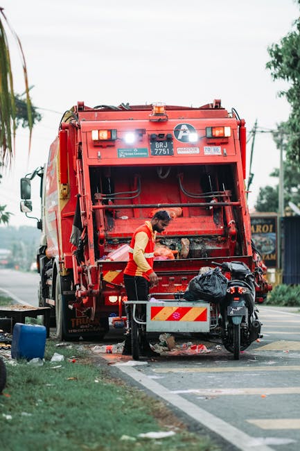 A waste collection worker is engaged in emptying a large red rubbish truck positioned on the side of a street, with its rear open revealing mechanical components and a compacted waste compartment. The worker, dressed in a yellow high-visibility vest and dark trousers, is standing next to a small black motorcycle parked close to the truck, with a black plastic bag placed on the motorcycle's side carrier. The street environment includes a paved shoulder, a grassy verge on the left, and a background featuring a small building or shop with signage, trees, and overhead utility wires, indicating a suburban or urban area. The scene is illuminated by natural daylight, with an overcast sky contributing to soft, diffused lighting. This image visually supports the context of private waste collection and on-site rubbish removal services performed by companies such as Rubbish Removal Fulham, highlighting the process of waste loading and disposal outside a residential or commercial property.