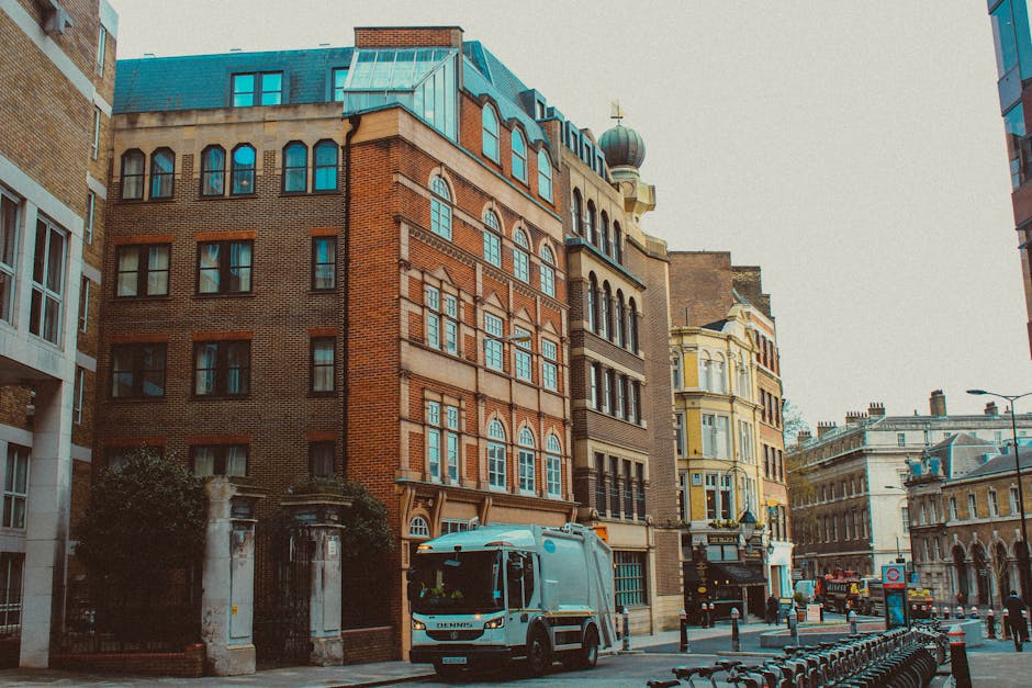 A city street scene featuring a large, multi-storey brick building with decorative architectural details and arched windows, situated among other historic and modern structures. In the foreground, a grey waste collection truck with the Rubbish Removal Fulham logo is parked on the pavement, adjacent to a row of black metal bike racks. The surrounding environment includes a small gated area with stone pillars and greenery at the building's entrance, and other vehicles and buildings can be seen further down the street. The sky appears overcast, providing soft, diffuse lighting over the scene. This image visually supports the context of private waste management and independent rubbish removal services, illustrating urban waste collection processes in a bustling city environment.