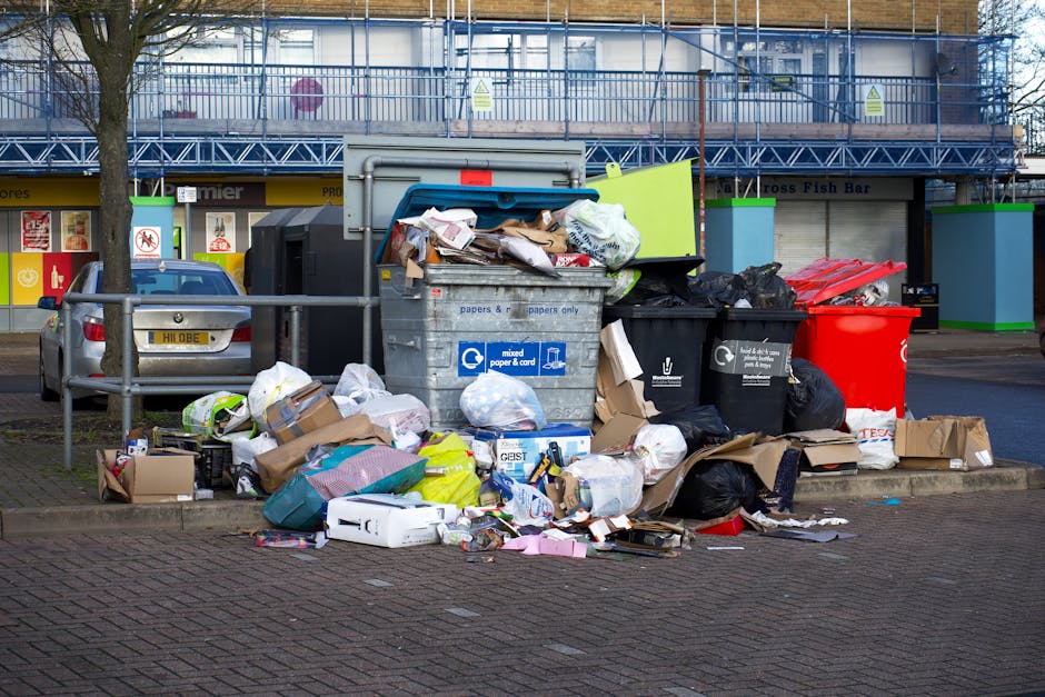 A waste collection worker is engaged in emptying a large red rubbish truck positioned on the side of a street, with its rear open revealing mechanical components and a compacted waste compartment. The worker, dressed in a yellow high-visibility vest and dark trousers, is standing next to a small black motorcycle parked close to the truck, with a black plastic bag placed on the motorcycle's side carrier. The street environment includes a paved shoulder, a grassy verge on the left, and a background featuring a small building or shop with signage, trees, and overhead utility wires, indicating a suburban or urban area. The scene is illuminated by natural daylight, with an overcast sky contributing to soft, diffused lighting. This image visually supports the context of private waste collection and on-site rubbish removal services performed by companies such as Rubbish Removal Fulham, highlighting the process of waste loading and disposal outside a residential or commercial property.